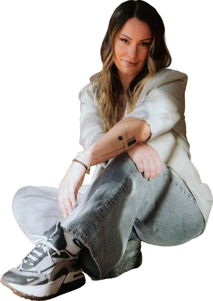Stephy Schutta sitzt in weißem Blazer, Jeans und Sneakern auf dem Boden